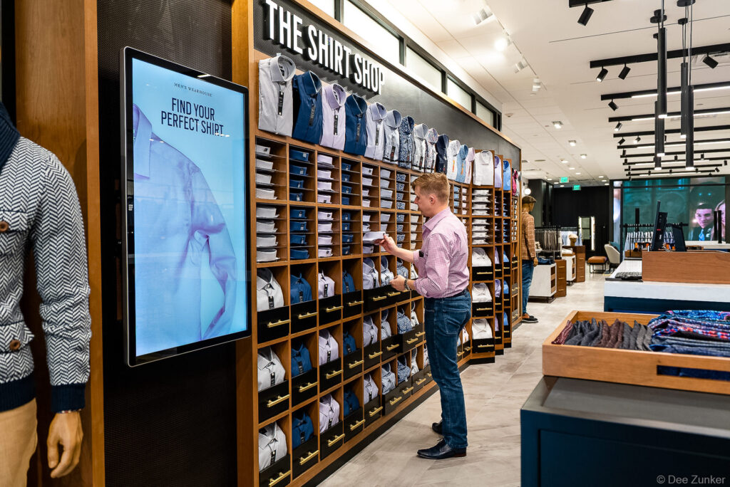 Customer browses folded dress shirts on wood cubby wall display inside Men's Wearhouse retail store, Houston commercial lifestyle branding photography.