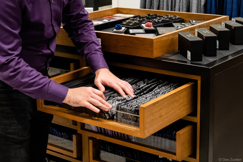 Sales associate in purple shirt browses neckties in wooden drawer display at Houston menswear retailer, with cufflinks and pocket squares above.