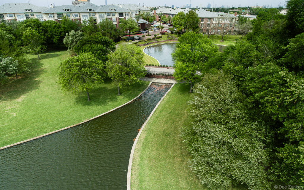 Aerial drone photo of Meyer Park Houston showing a winding canal, lush green lawns, mature trees, and residential buildings beyond.