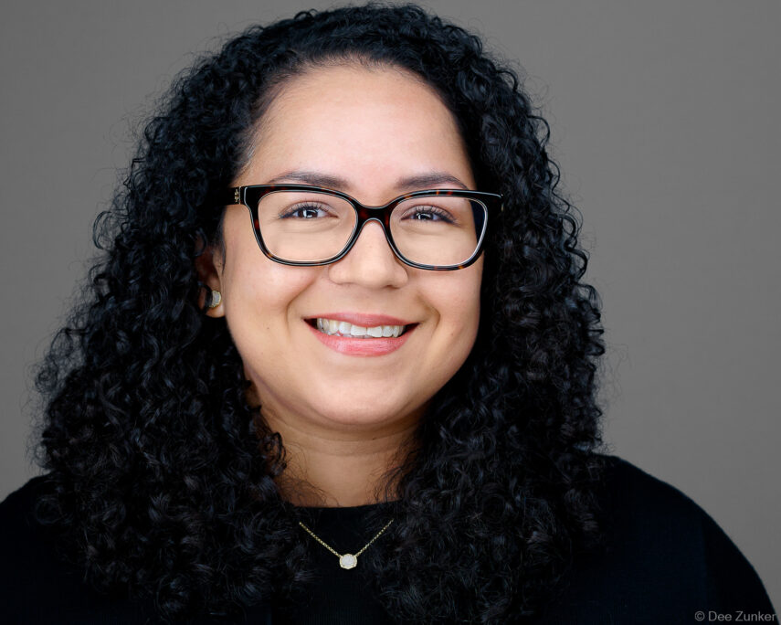 Professional corporate headshot of a smiling woman with curly hair and glasses, studio portrait taken for Bellaire TX office headshots