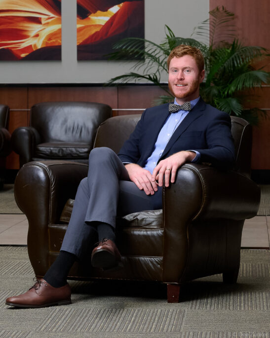 Male professional in navy blazer and bow tie seated in leather chair during corporate staff portrait session at OMS Houston.