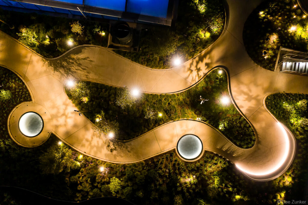 Overhead drone view of the POST Houston rooftop garden at night. Illuminated pathways and glowing glass skylights are visible.