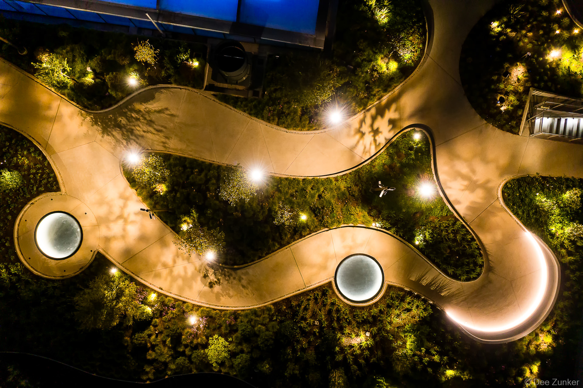 Overhead drone view of the POST Houston rooftop garden at night. Illuminated pathways and glowing glass skylights are visible.