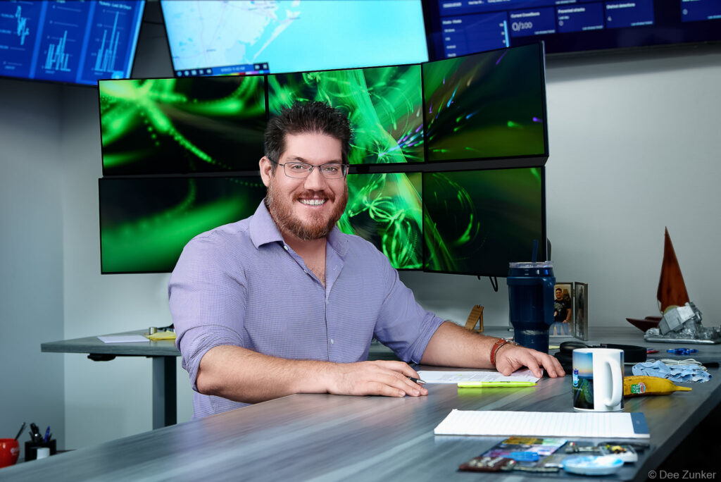 Male executive smiles at a standing desk surrounded by multiple monitors displaying data dashboards, Houston corporate branding portrait by Dee Zunker Photography.
