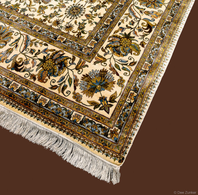 Close-up product photo of an antique silk oriental rug with ivory field, floral motifs, gold border, and cream fringe.