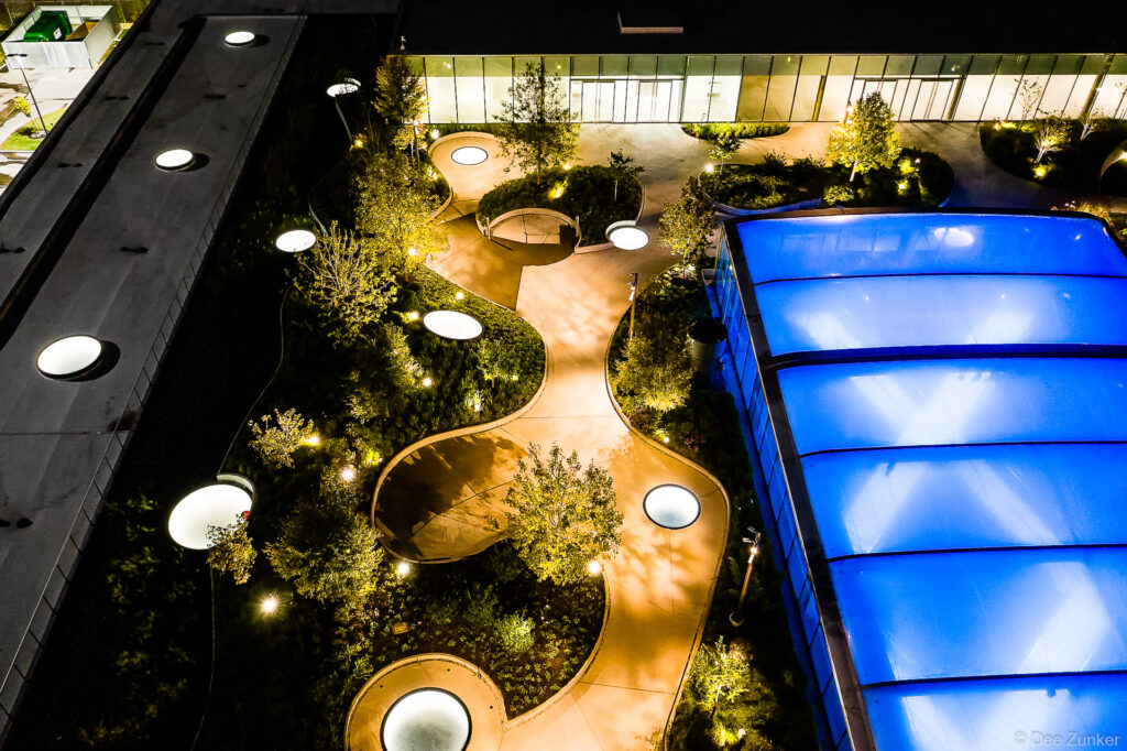 Aerial night view of Post Houston rooftop garden with illuminated curved pathways, glowing skylights, trees, and blue glass canopy.