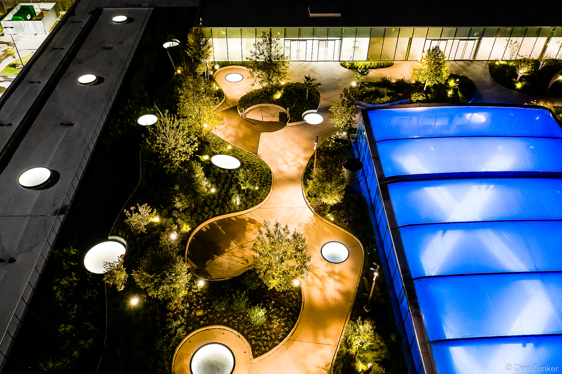 Aerial night view of Post Houston rooftop garden with illuminated curved pathways, glowing skylights, trees, and blue glass canopy.