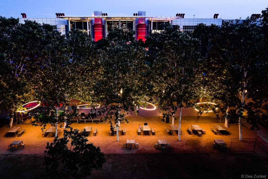 Aerial view of Discovery Green park in Houston at dusk with illuminated tables, trees, and red-lit building facade.