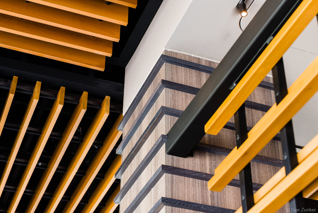 Close-up architectural product photo showing golden wood batten ceiling panels and dark-trimmed staircase in a modern commercial interior.