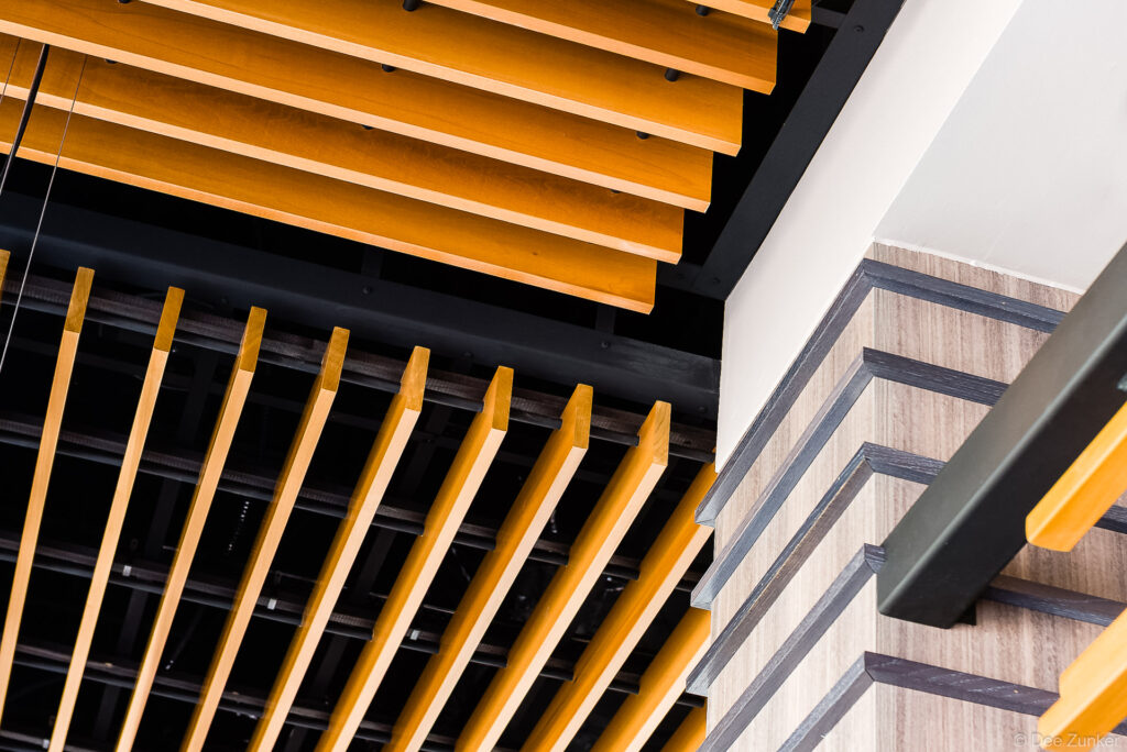 Close-up of warm wood ceiling baffles and slatted panels beside dark-trimmed stairs in a modern commercial interior Houston.