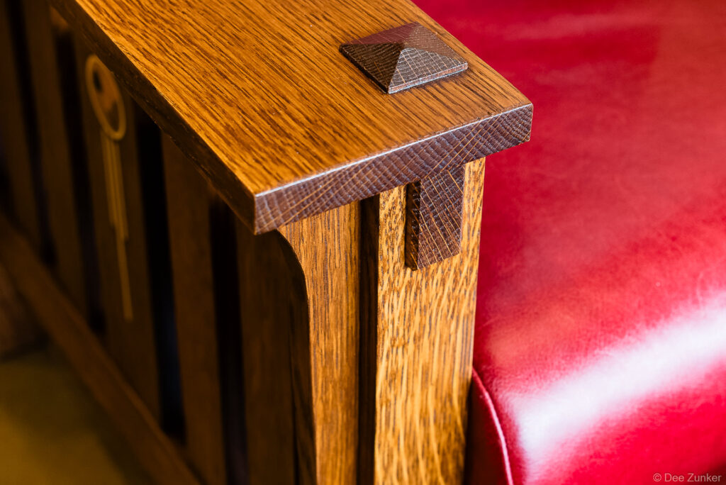 Close-up product photo of a quarter-sawn oak Mission style chair arm with pyramid cap detail and red leather seat cushion.