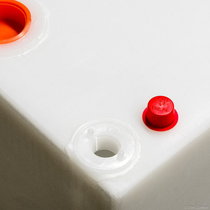 Close-up product photo of white polyethylene boat storage box showing threaded port and red plastic plug caps on gray background.