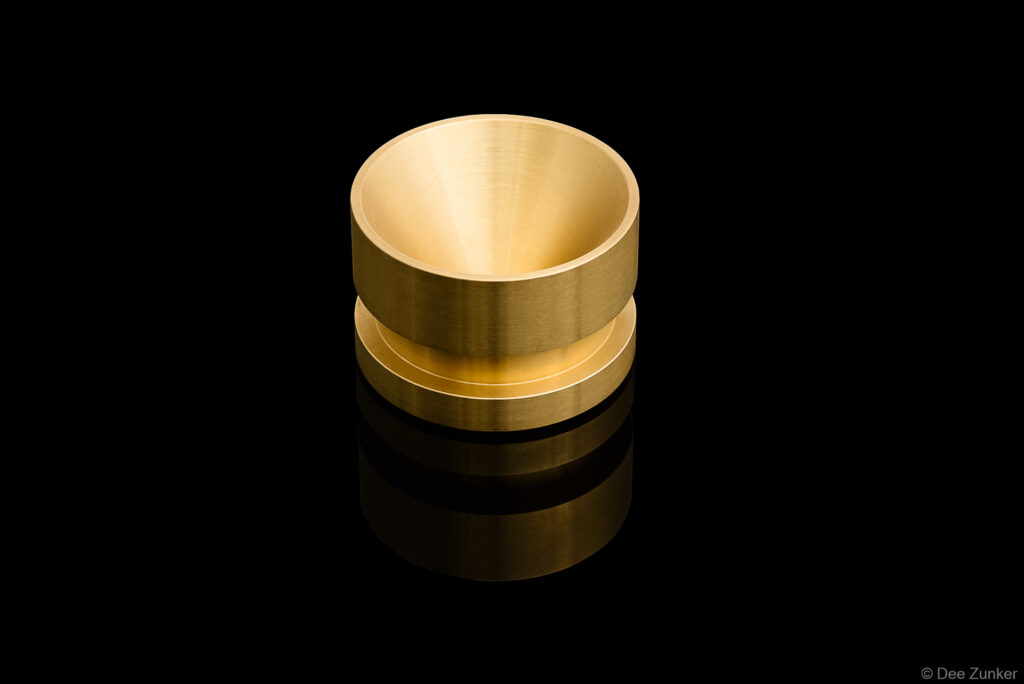 Machined brass cylindrical cup component photographed on reflective black background, Houston industrial product photography by Dee Zunker.