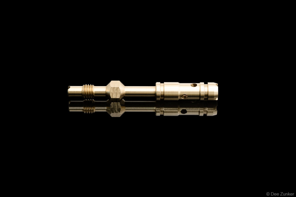Polished brass industrial drilling tool component with threaded fitting and ported cylinder, photographed on reflective black background.