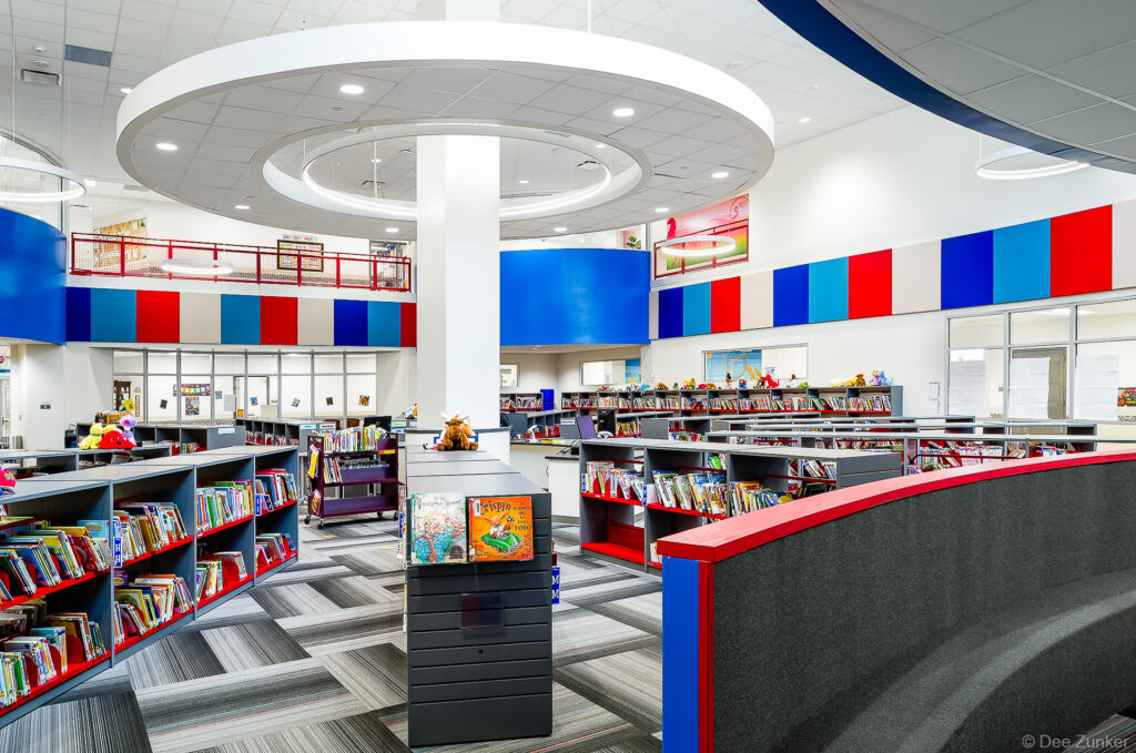 Colorful elementary school library interior featuring gray and red bookshelves, acoustic ceiling panels in blue and red, Houston TX.