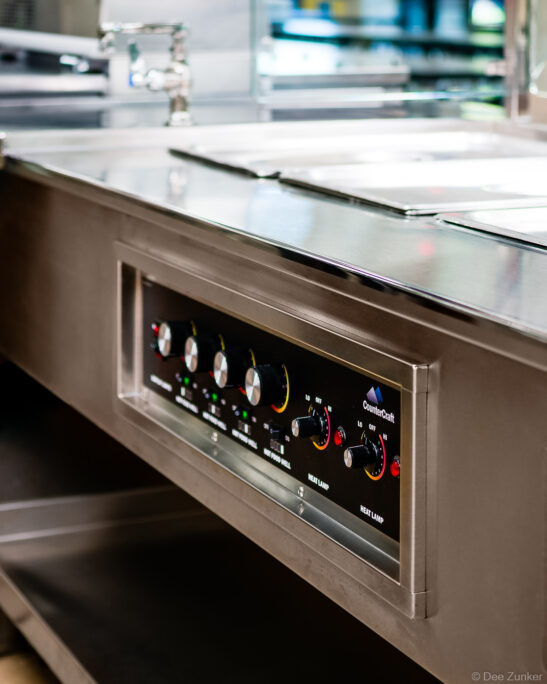 Close-up of stainless steel institutional food service counter with CounterCraft heat lamp and hot food well controls, Houston commercial product photography.
