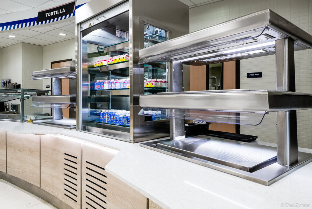Stainless steel cafeteria serving counters with refrigerated milk display cases and heated food wells, institutional food service equipment photography.