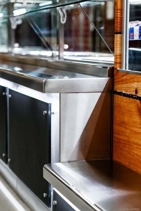 Close-up of brushed stainless steel food service counter with glass sneeze guard and wood panel accent, Houston commercial product photography.