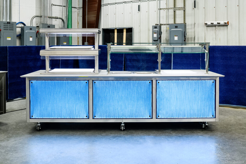 Stainless steel institutional food service counter with blue LED panels, sneeze guards, and heat lamps, photographed for Houston product portfolio.
