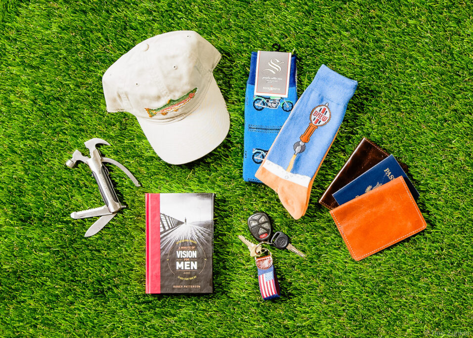 Flat lay product photo of men's gifts including a fishing cap, novelty socks, multi-tool, book, car keys, and leather passport wallet on green turf.