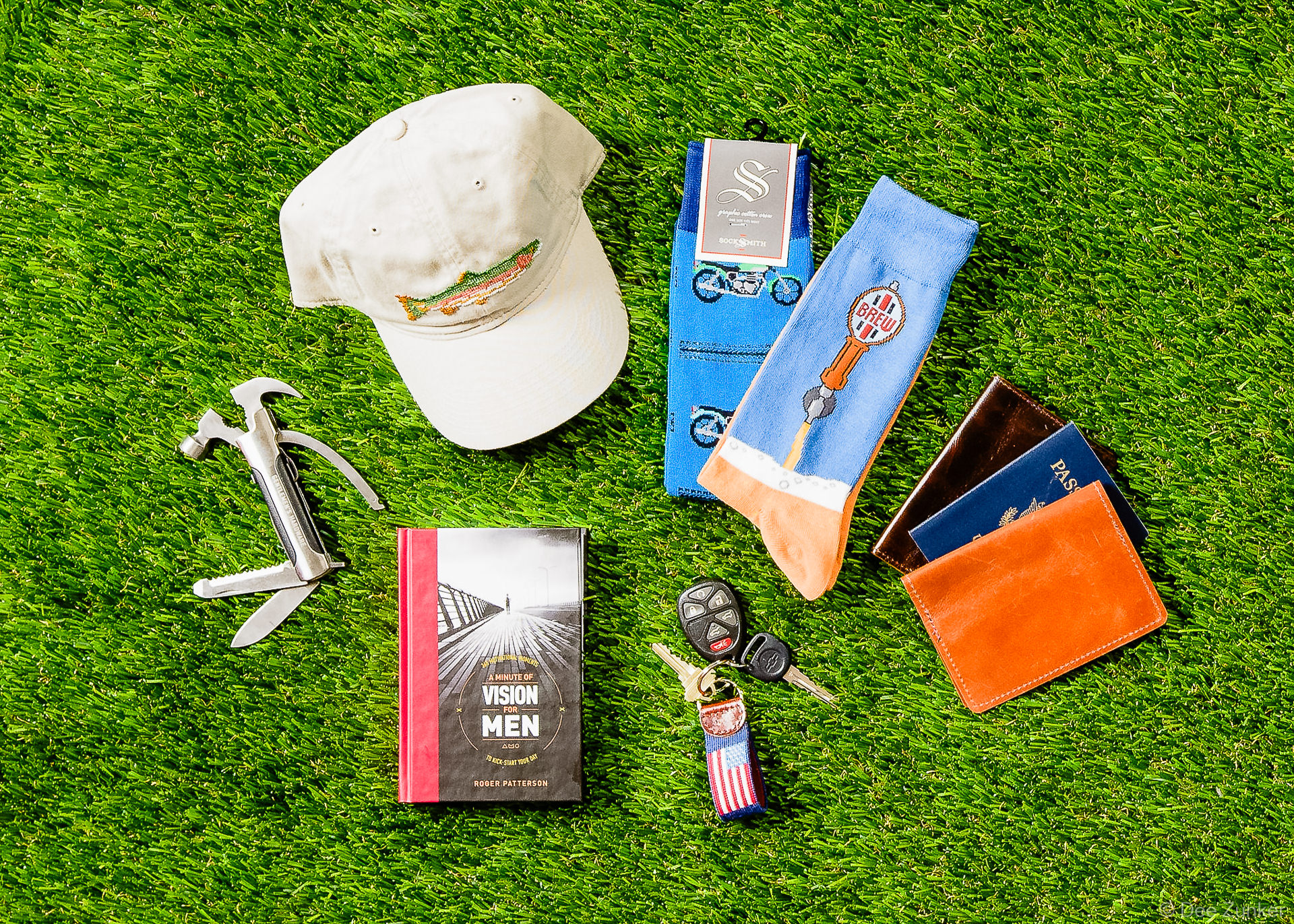 Flat lay product photo of men's gifts including a fishing cap, novelty socks, multi-tool, book, car keys, and leather passport wallet on green turf.