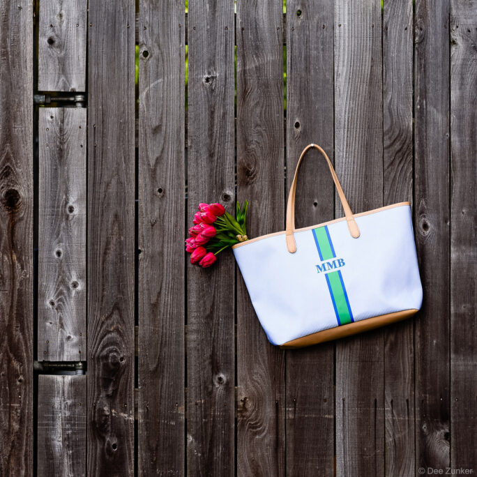 White monogrammed MMB tote bag with green and blue stripes and tan leather handles displayed with red tulips against a wooden fence.