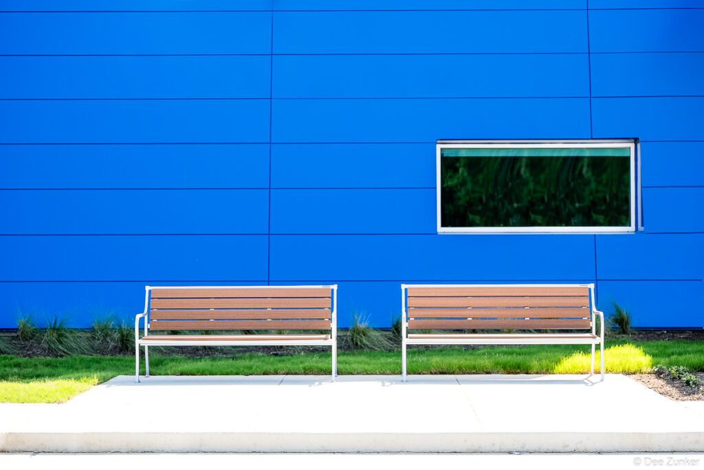 Two wood-slat benches with white metal frames against a bold blue paneled institutional building exterior, Houston commercial photography.