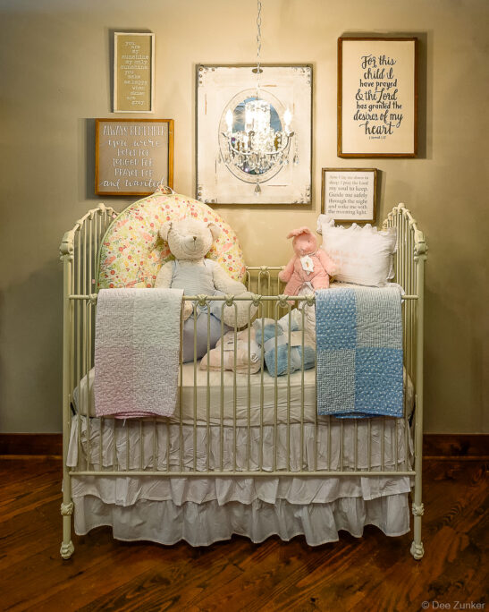 Styled baby nursery vignette featuring a white iron crib, plush toys, quilts, and inspirational wall art for retail gift product photography.