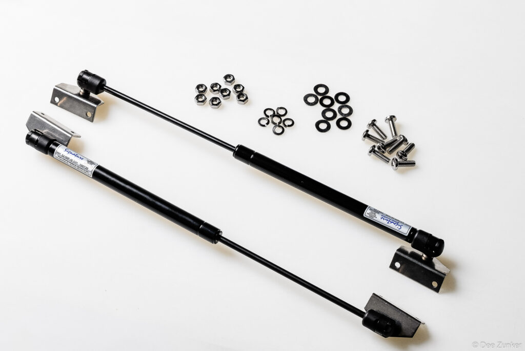 Two black gas struts with mounting brackets, nuts, washers, and screws arranged on white background for industrial catalog photography.