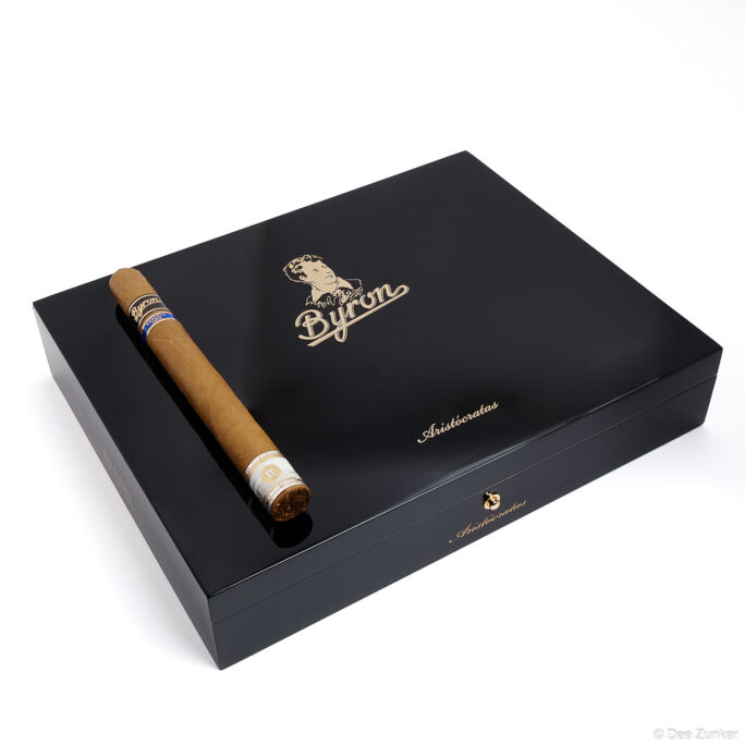 Byron Aristocratas premium cigar in front of glossy black humidor box, retail product photography on white background by Houston studio.