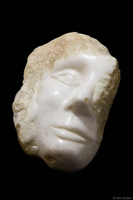 Close-up product photo of a white marble ancient face sculpture fragment against black background, Houston studio photography.
