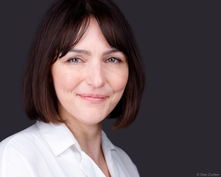 Woman with dark bob hair smiles confidently in a studio corporate headshot against a gray background, Austin professional photography.