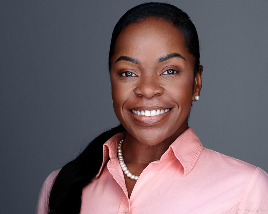 Smiling professional woman in pink blouse and pearl necklace poses for a corporate studio headshot against a gray background in Houston.