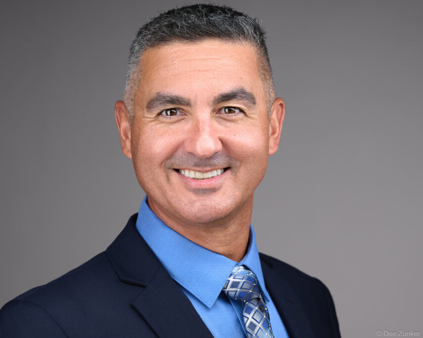Male executive smiles in studio corporate headshot, wearing navy suit and blue tie against gray background, Houston professional photography.