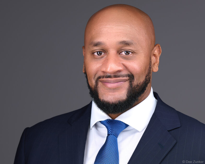 Bald African American male professional in navy suit and blue tie smiles confidently in a Houston studio headshot against a gray background.