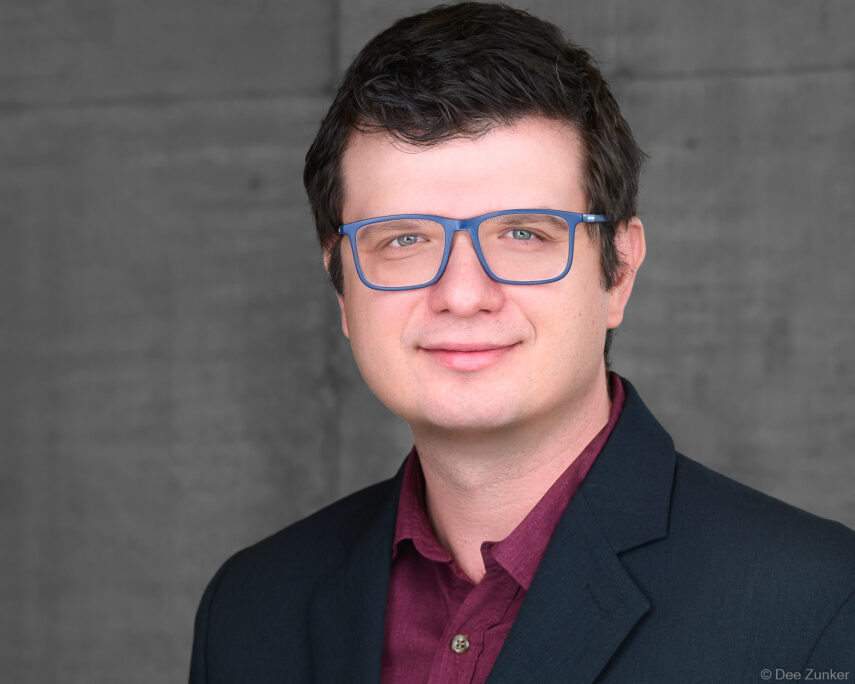 Male professional in blue-framed glasses and dark blazer poses for a corporate studio headshot against a gray background in Houston.