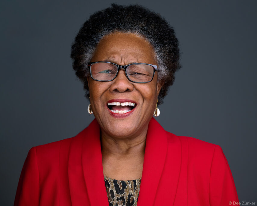 Smiling older woman in red blazer and glasses laughs during professional studio headshot session against gray background in Houston.