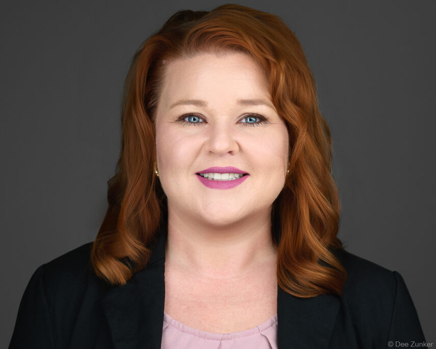 Red-haired professional woman with blue eyes smiles in a studio corporate headshot against a gray background, Houston TX.