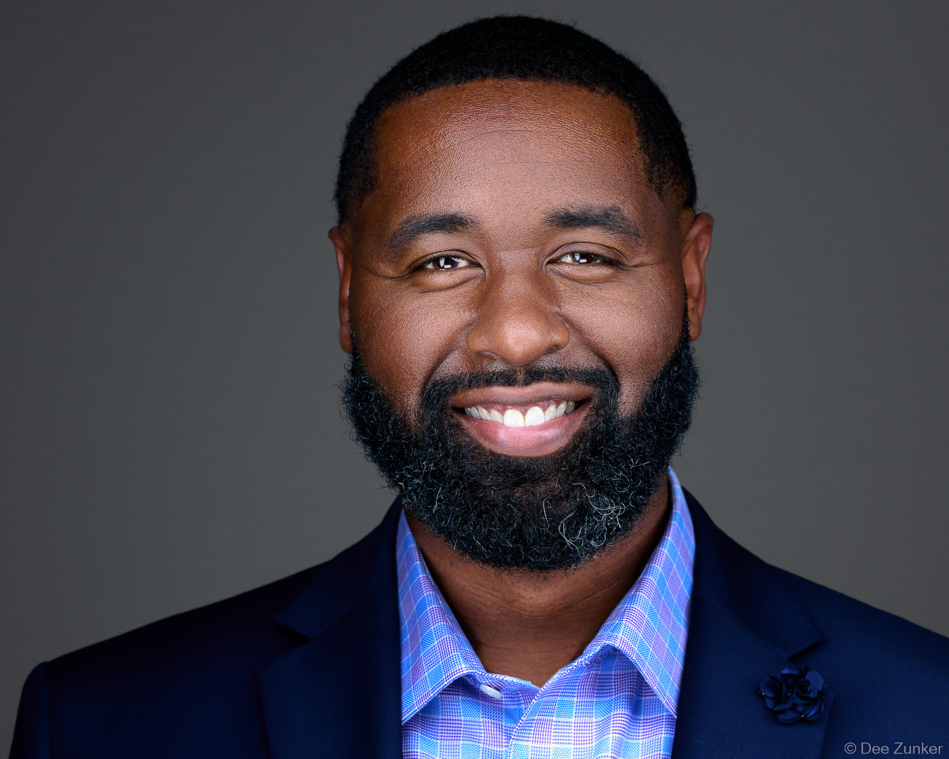 Smiling professional man in navy blazer and plaid shirt poses for a professional studio headshot against a gray background in Houston.