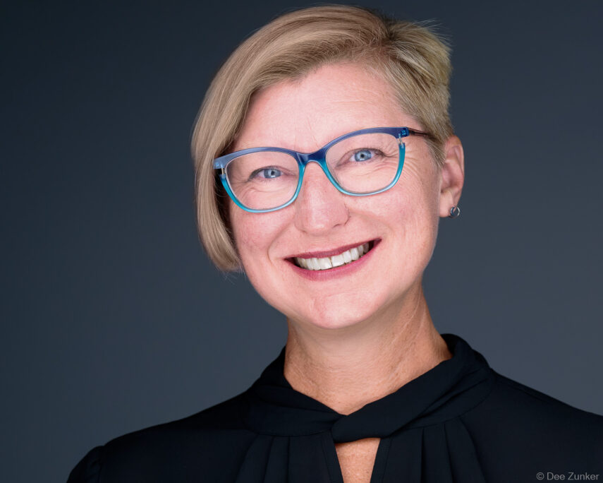 Smiling professional woman with blue glasses and short blonde hair poses for a corporate studio headshot against a gray background in Houston.