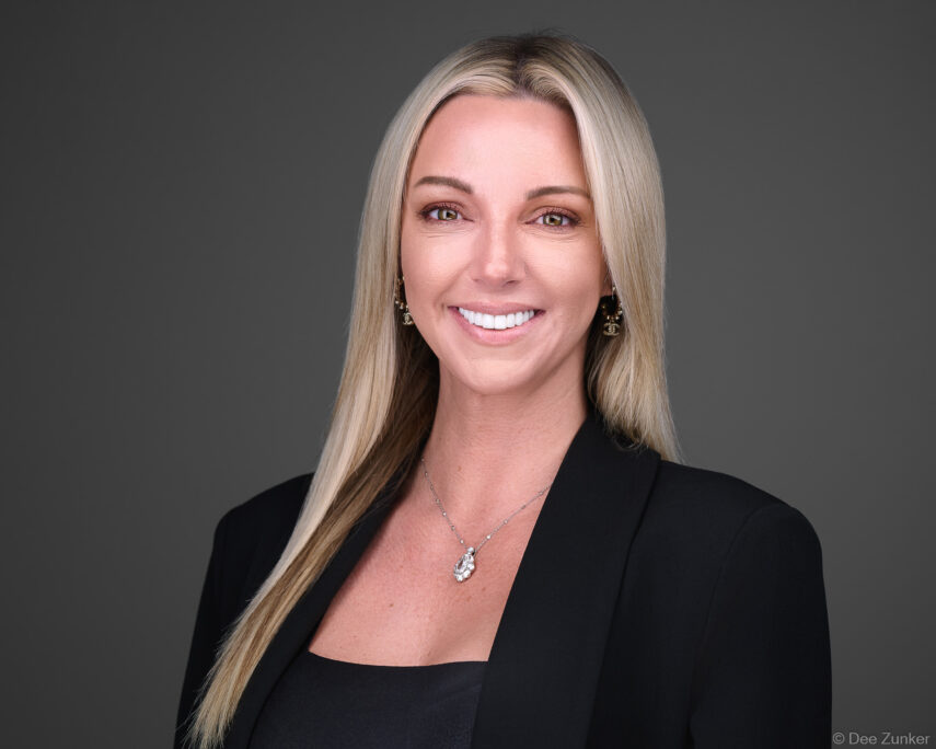 Blonde female executive smiles in black blazer against gray studio backdrop, professional corporate headshot, Houston, TX.