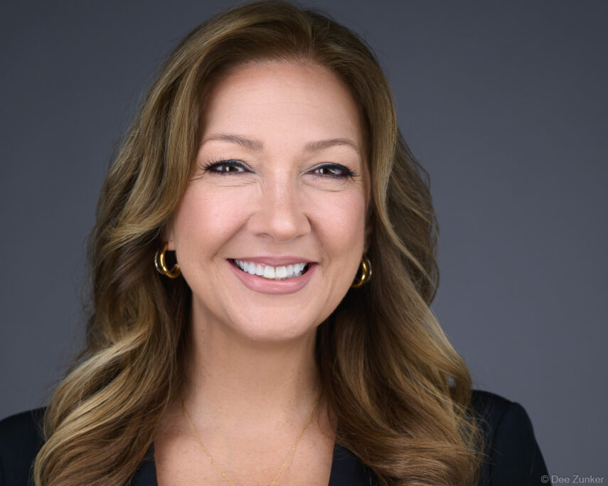 Professional corporate headshot of a woman with wavy blonde-brown hair, gold hoop earrings, and a black blazer, photographed on a gray studio background in Houston.