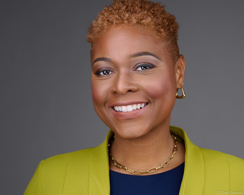 Smiling professional woman in chartreuse blazer and gold chain necklace poses for a corporate studio headshot against a gray background in Houston.