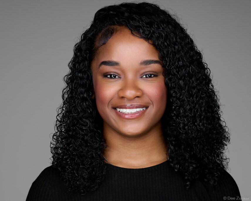Young professional woman with curly hair smiles in a studio corporate headshot against a gray background in Houston.