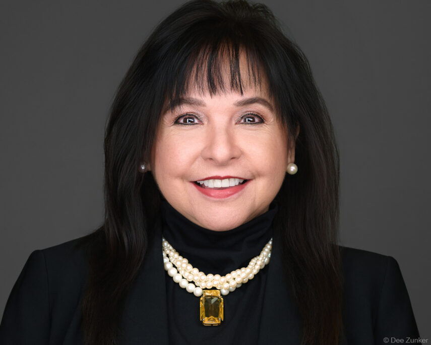 Female executive smiling in studio headshot, wearing pearl necklace and black blazer, photographed against a gray background in Houston.