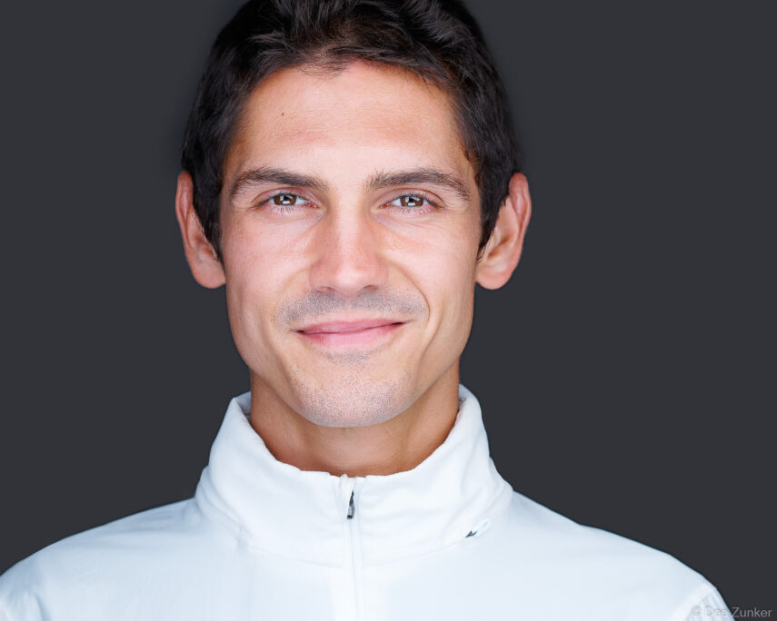 Male professional smiling in a studio corporate headshot, wearing a white zip-neck jacket against a dark gray background, Houston.