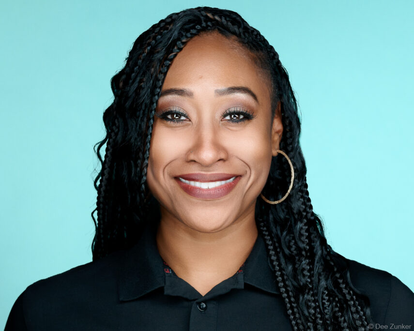 Woman with box braids smiles in a professional corporate headshot, wearing a black polo shirt against a teal studio background, Houston.