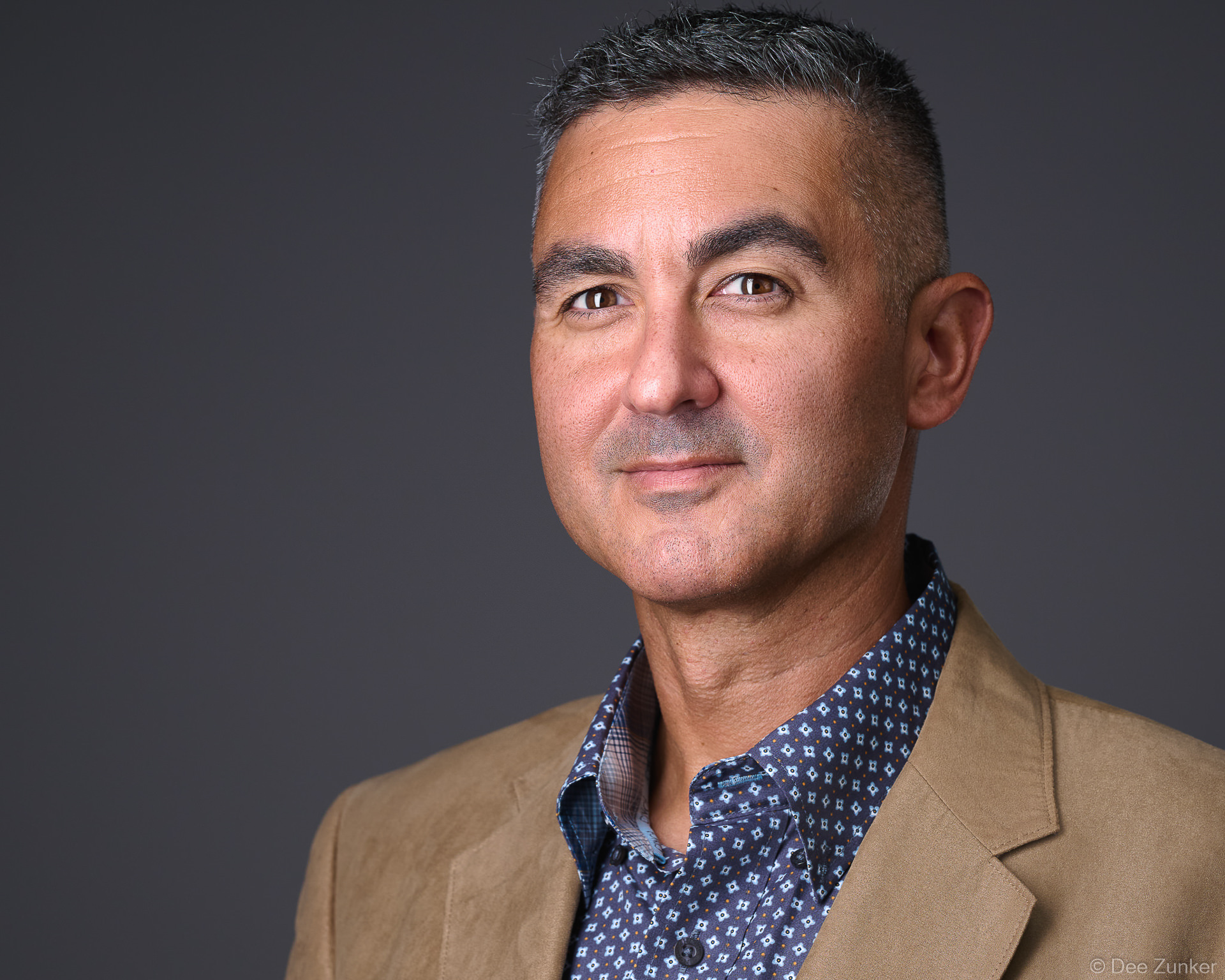 Middle-aged male professional in tan blazer and patterned shirt poses for a corporate studio headshot against a gray background in Houston.
