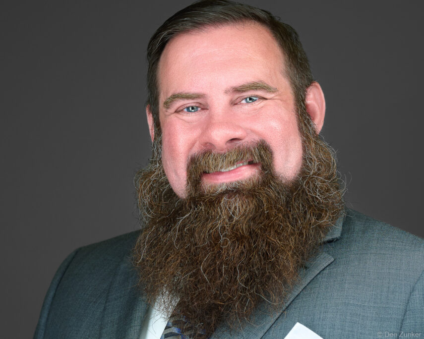 Male professional with full brown beard and blue eyes in gray suit, studio corporate headshot against gray background, Houston TX