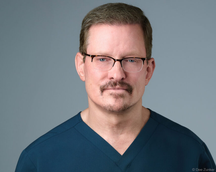 Male healthcare professional in navy scrubs and tortoiseshell glasses, studio corporate headshot against gray background, Houston TX